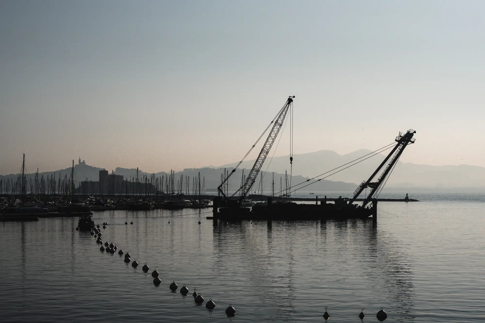 A crane barge moored in a harbour at dawn, with a line of buoys in the foreground