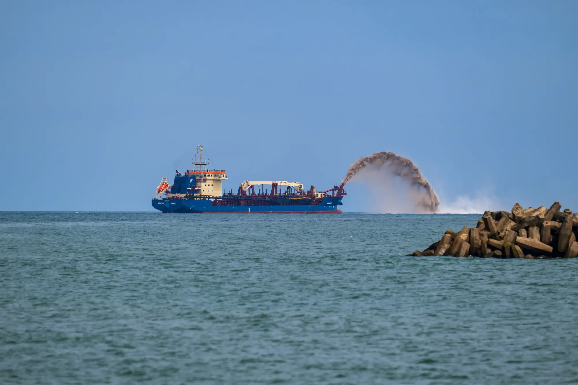A dredger spraying sand in an arc offshore, beyond a breakwater of concrete blocks